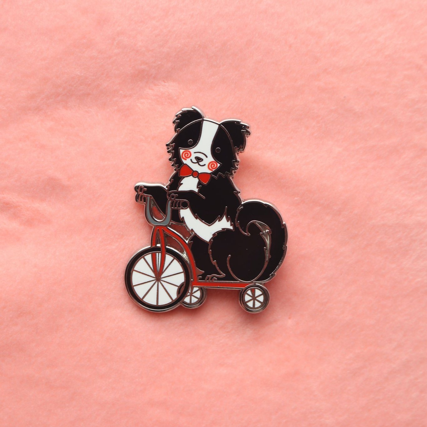 Enamel pin of a Black and White Border Collie wearing a red bow tie and painted red swirls on its cheeks. It is riding on a red tricycle.