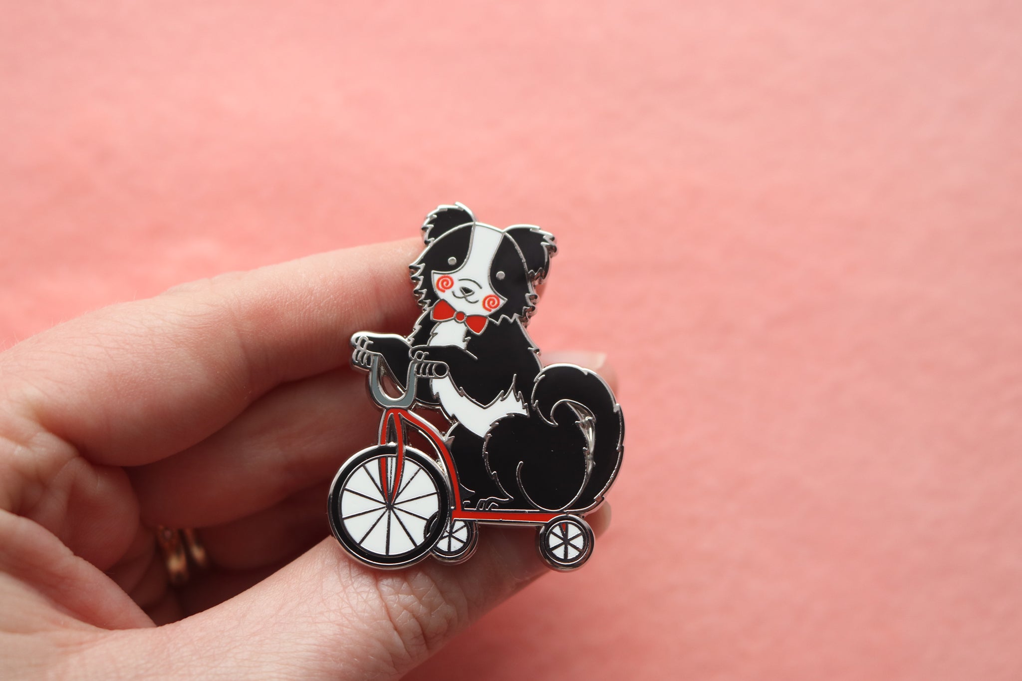 Hand holding an enamel pin of a Black and White Border Collie wearing a red bow tie and painted red swirls on its cheeks. It is riding on a red tricycle.