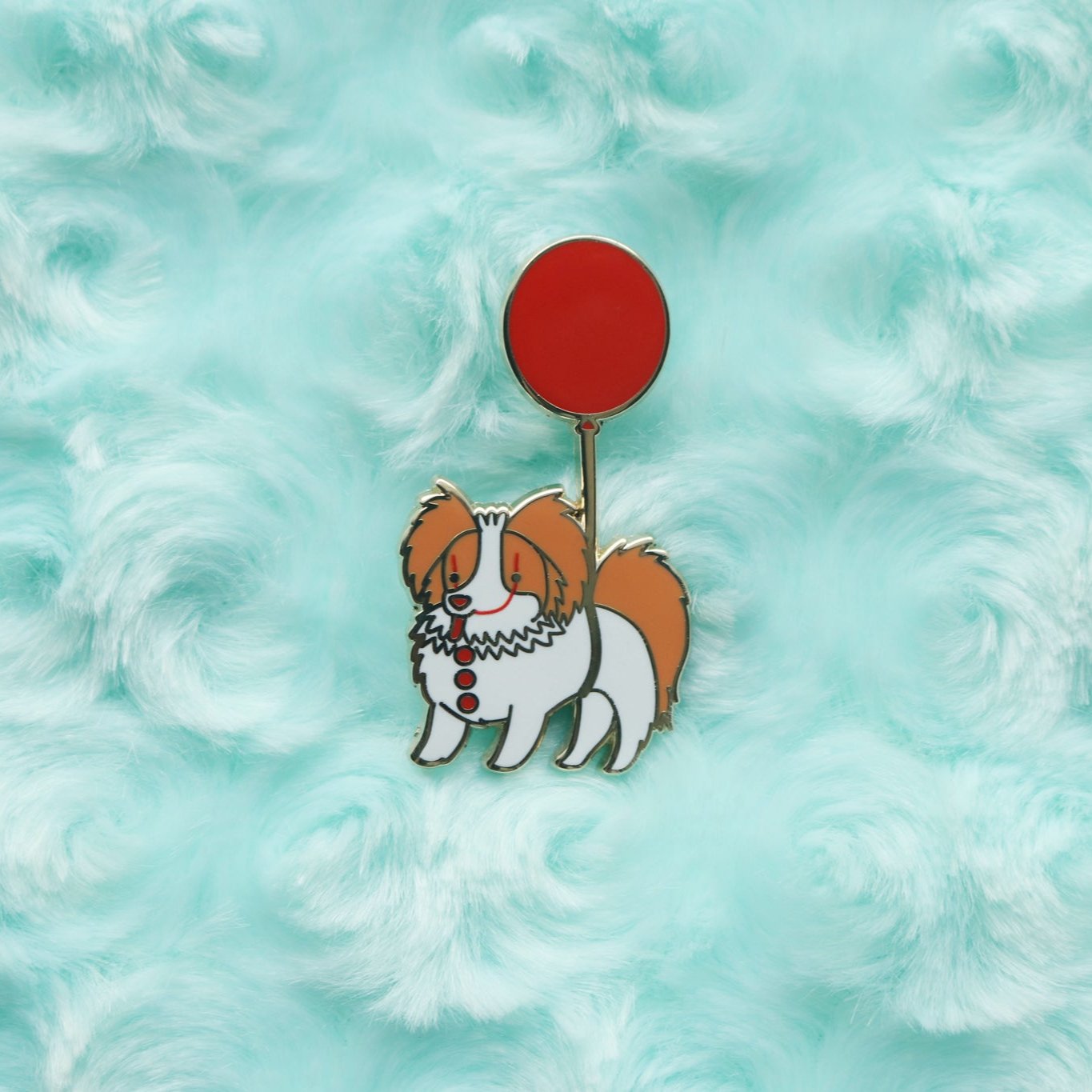 Enamel pin of an orange and white Papillon being carried by a red balloon. Dog has clown makeup on and three red buttons down its chest.