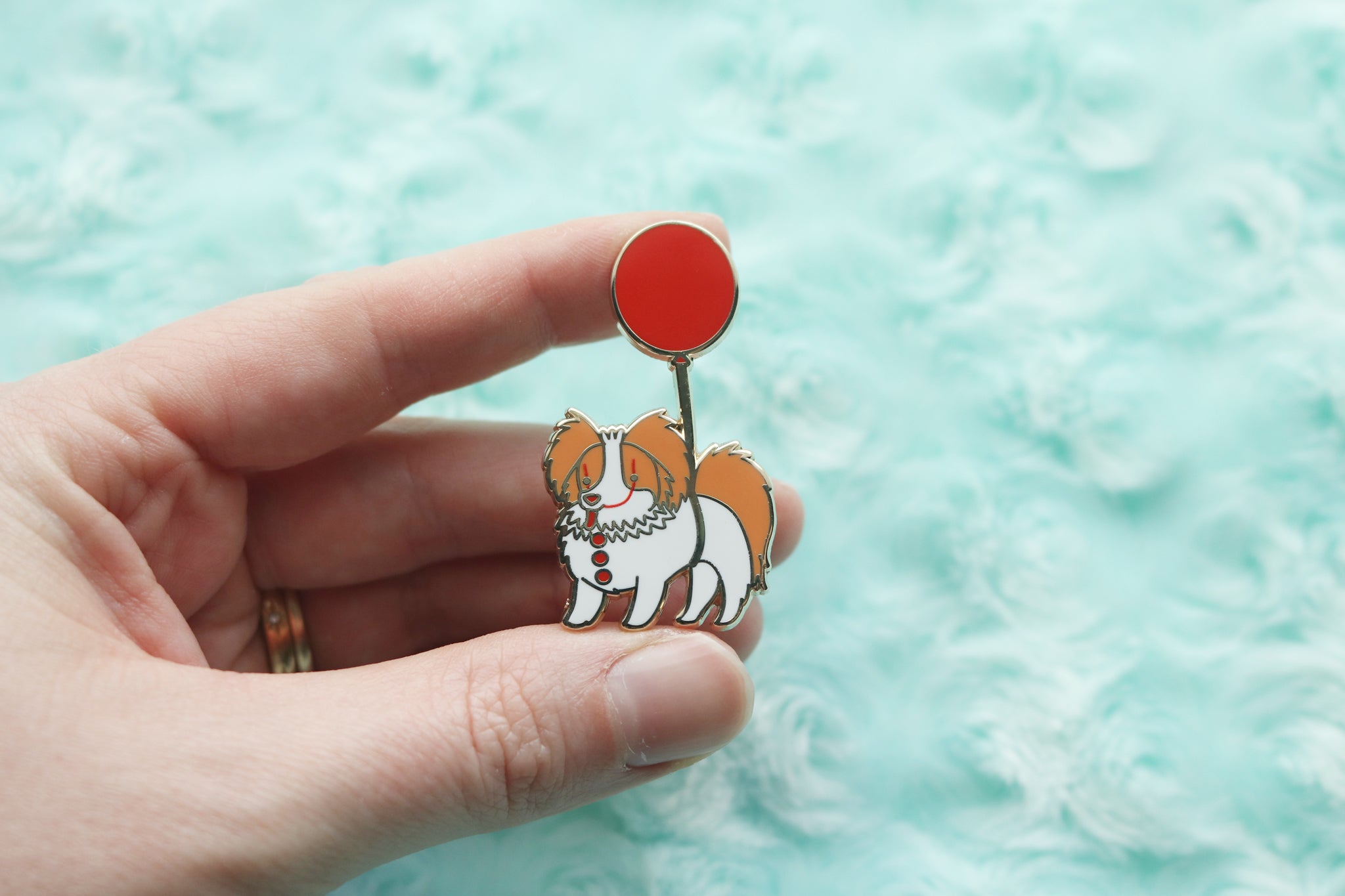 Hand holding an enamel pin of an orange and white Papillon being carried by a red balloon. Dog has clown makeup on and three red buttons down its chest.