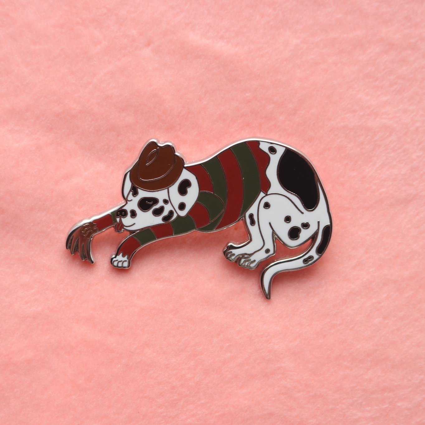 Enamel pin of a sleeping dalmatian wearing a green and red striped shirt, brown fedora, and a metal-clawed leather glove.