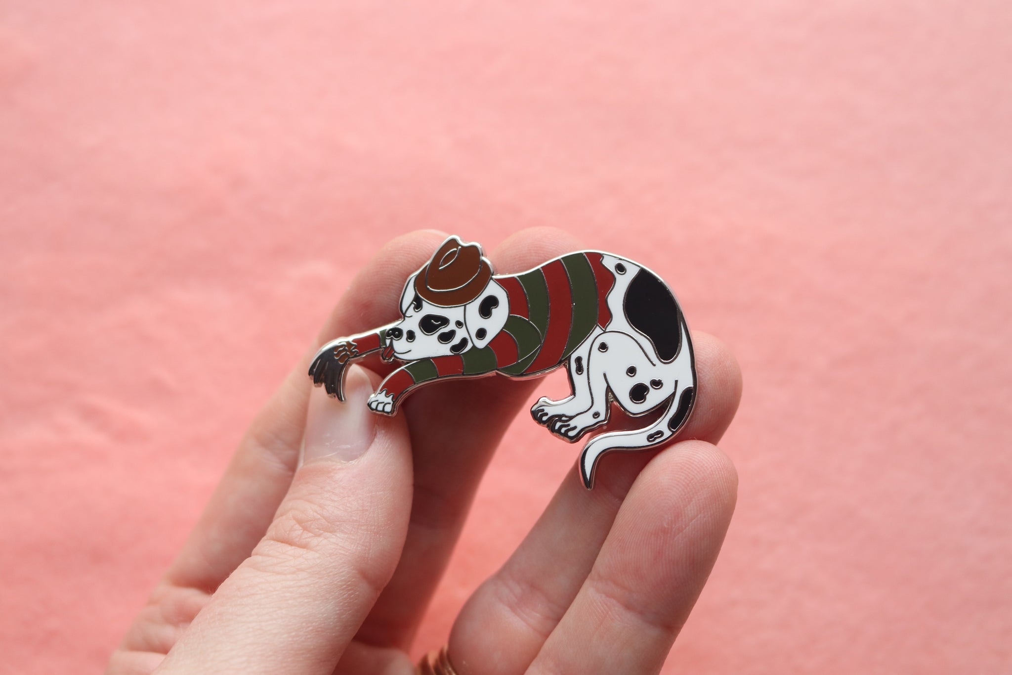 Hand holding an enamel pin of a sleeping dalmatian wearing a green and red striped shirt, brown fedora, and a metal-clawed leather glove.