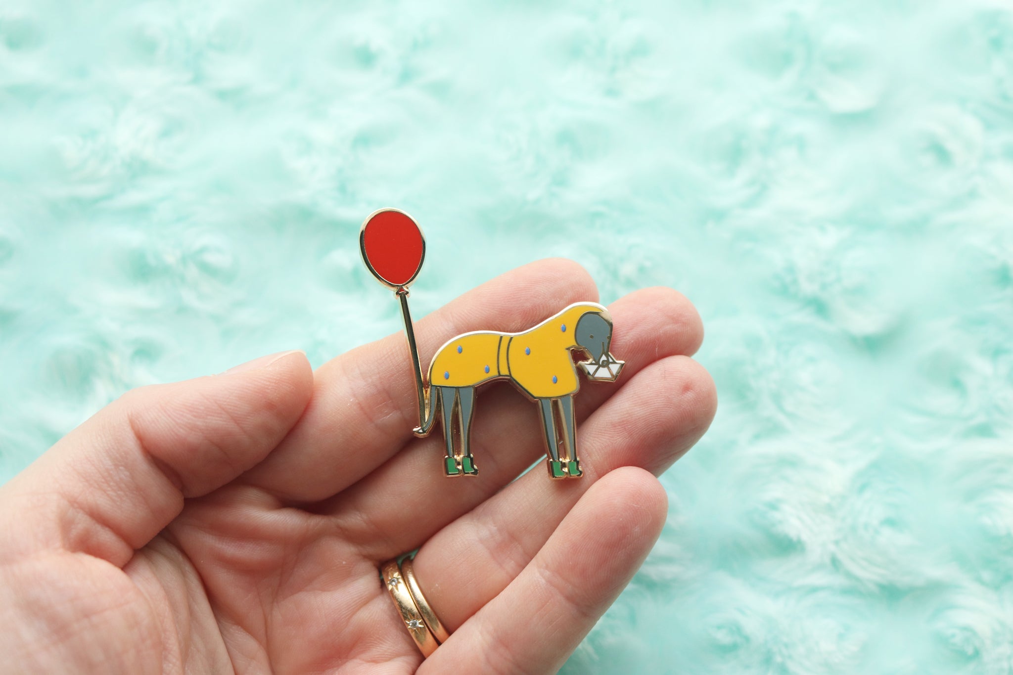 Hand holding an enamel pin of an italian greyhound wearing a yellow raincoat and green rain boots covered in rain. Dog holds a paper boat in it's mouth and has a red balloon tied to its tail.