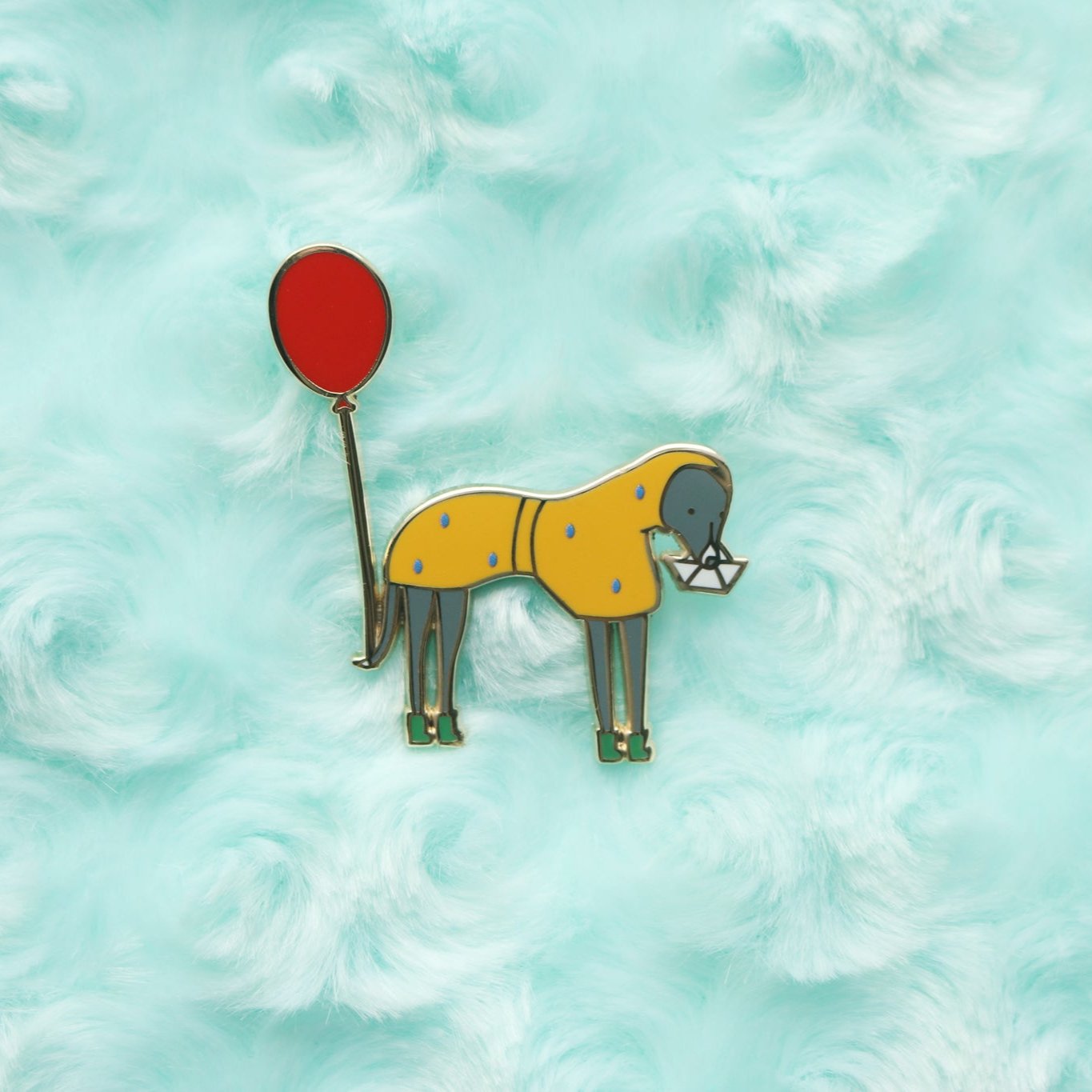 Enamel pin of an italian greyhound wearing a yellow raincoat and green rain boots covered in rain. Dog holds a paper boat in it's mouth and has a red balloon tied to its tail. 