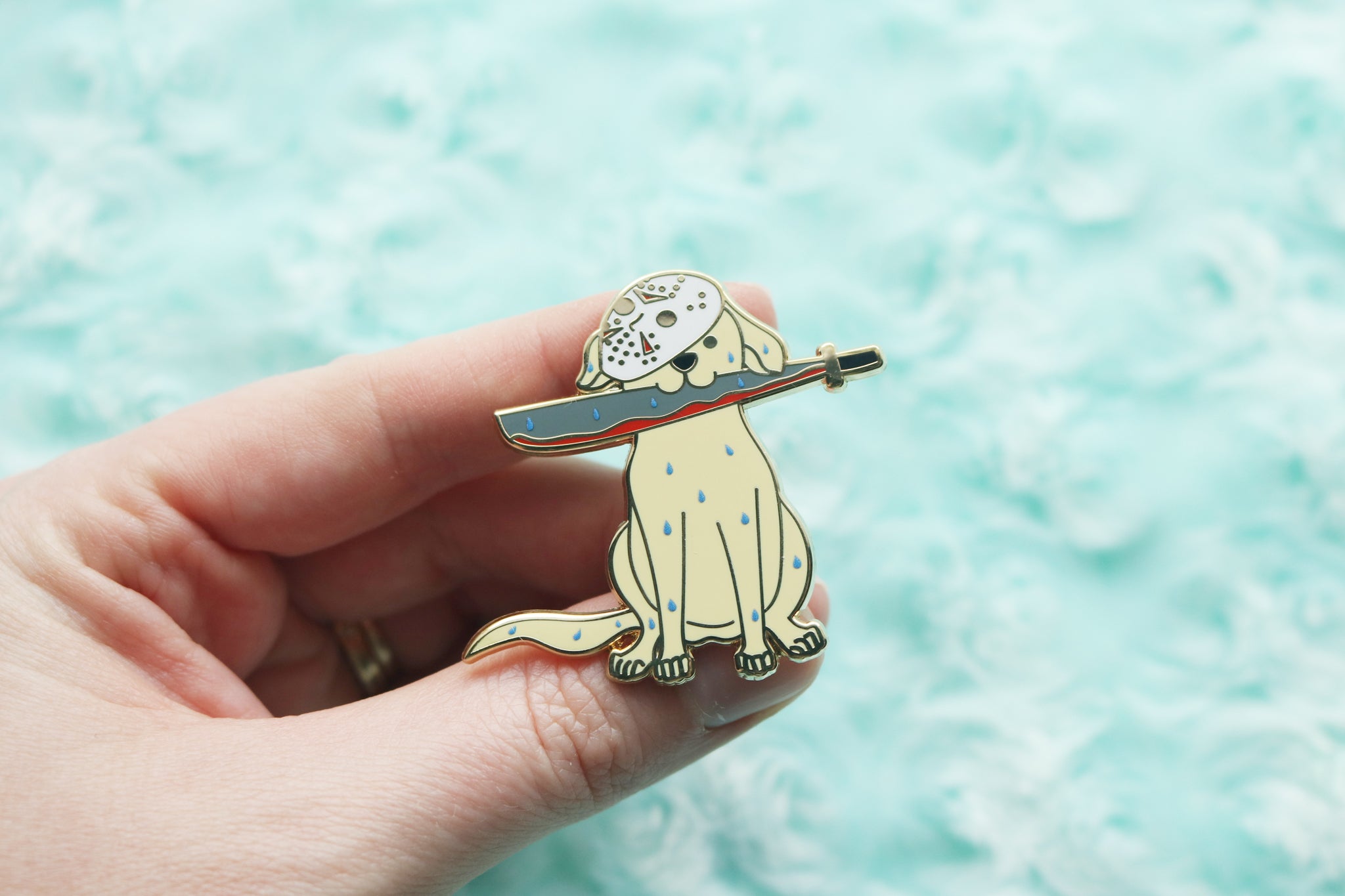 Hand holding an enamel pin depicting a wet Yellow Labrador Retriever holding a bloody machete. A hockey mask covers half of it's face.