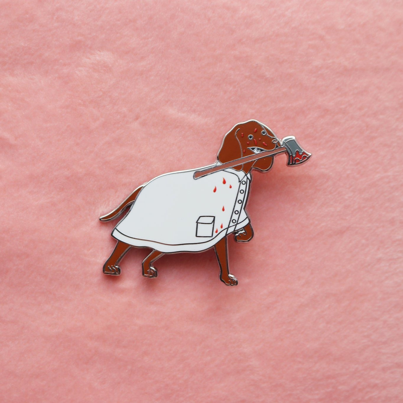 Enamel pin of a Vizsla wearing a bloody white raincoat and holding a bloody axe in its mouth.