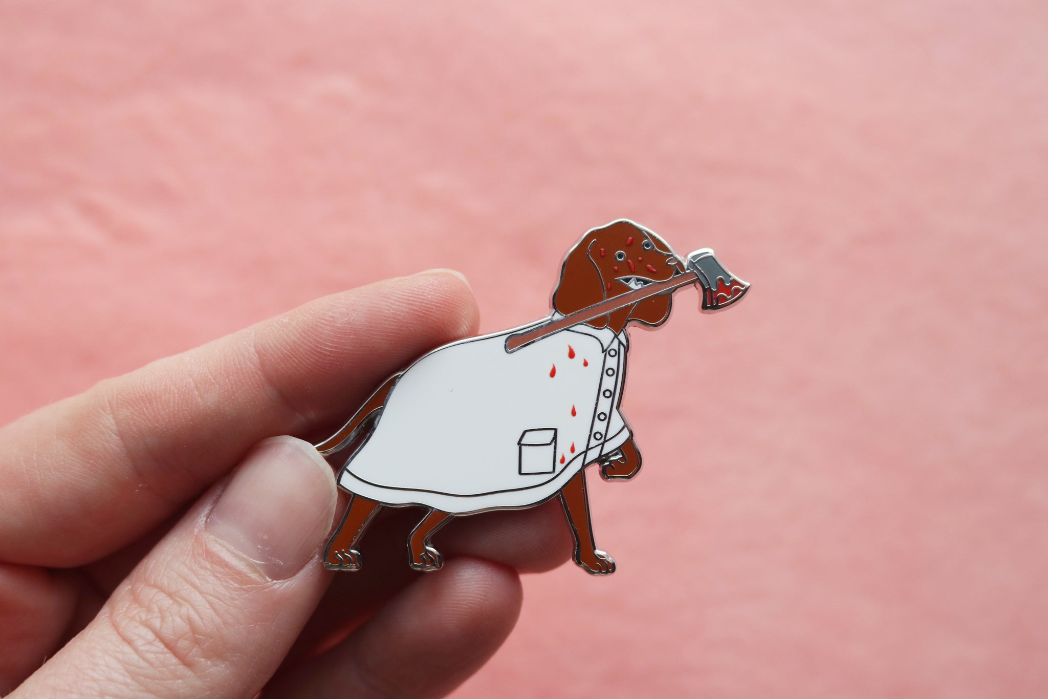 Hand holding an enamel pin of a Vizsla wearing a bloody white raincoat and holding a bloody axe in its mouth.