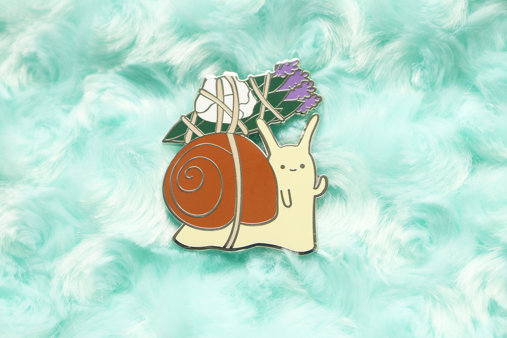 Enamel pin of a snail with a brown shell and beige body carrying a satchel of sage, rose petals, and lavender.