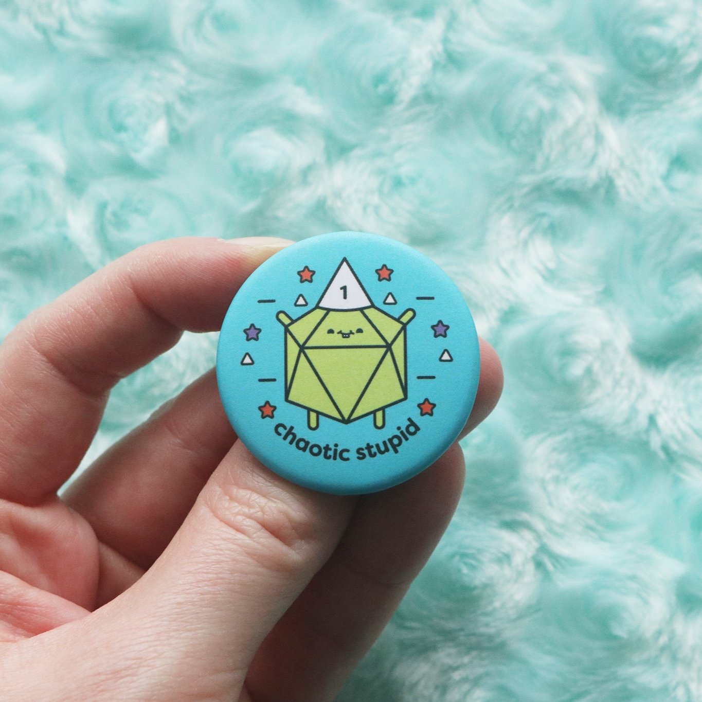 Hand holding a button that reads "Chaotic Stupid." A lime green d20 wears a white dunce cap with the number 1 on it.