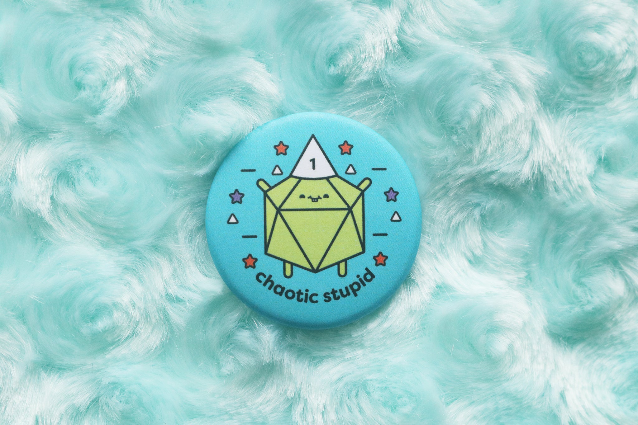 Button photographed onblue fur background. Button reads "Chaotic Stupid." A lime green d20 wears a white dunce cap with the number 1 on it.