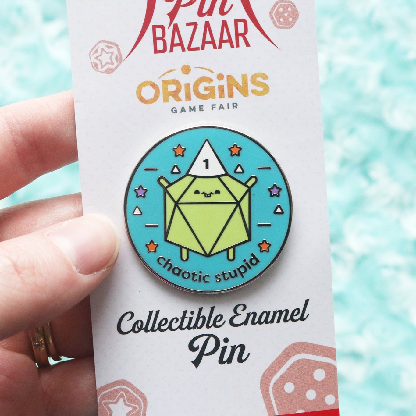 Hand holding a pin that reads "Chaotic Stupid" on a backer card that reads "Pin Bazaar, Origins Game Fair, Collectible Enamel Pin"