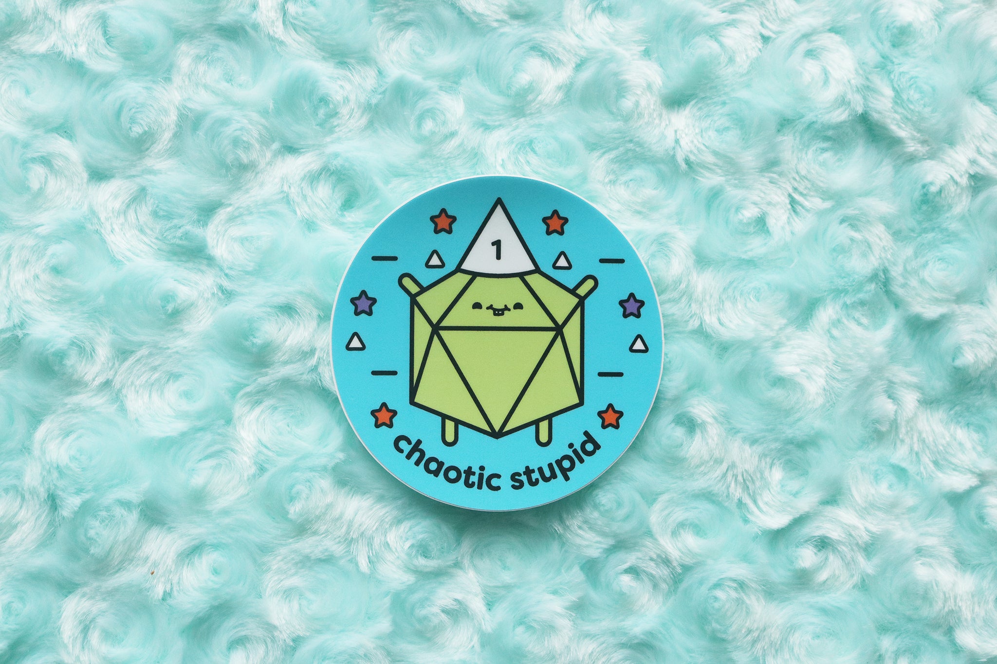 Sticker photographed on blue fur background. Sticker reads "Chaotic Stupid." A lime green d20 wears a white dunce cap with the number 1 on it.