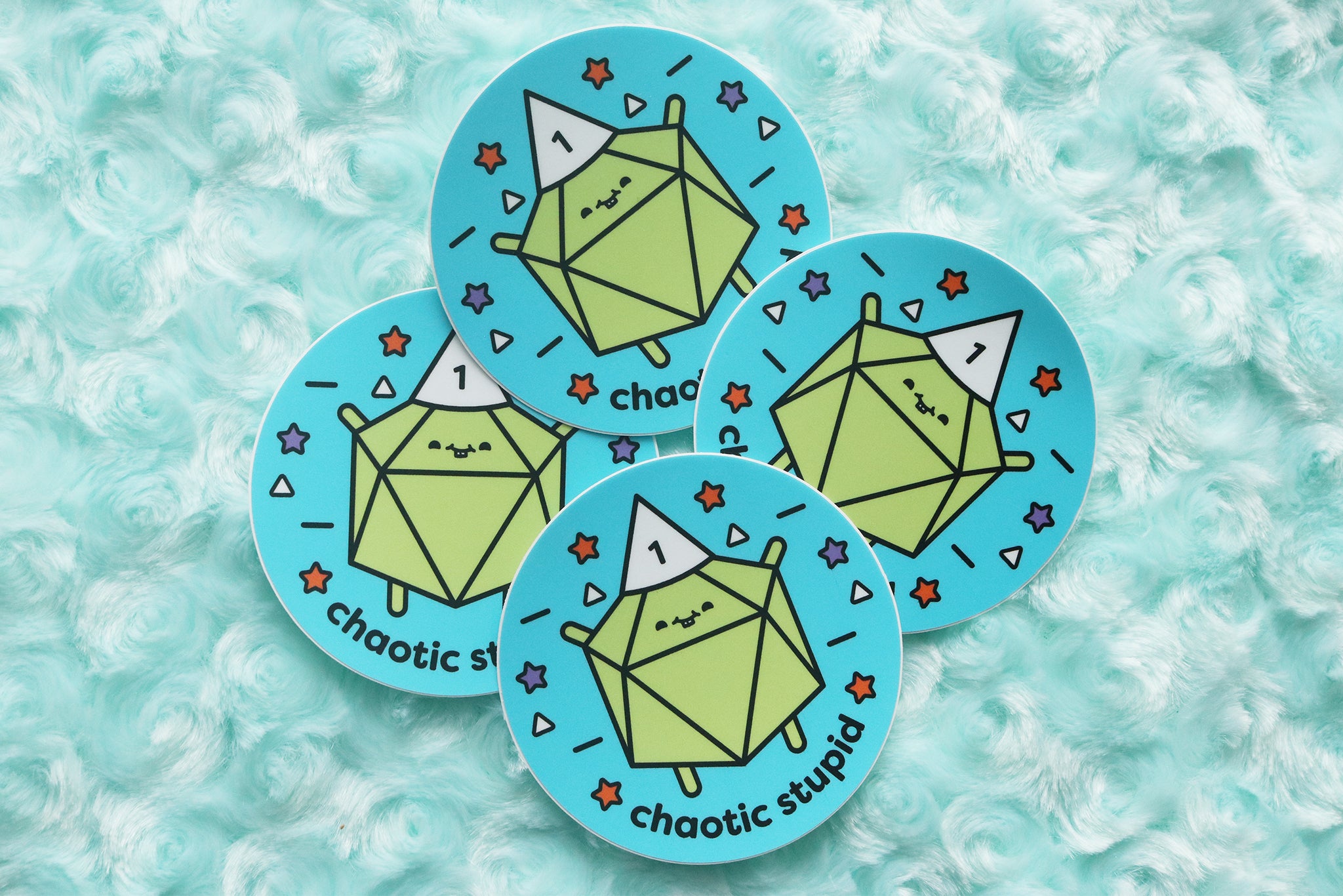 Pile of stickers photographed on blue fur background. Stickers reads "Chaotic Stupid." A lime green d20 wears a white dunce cap with the number 1 on it.