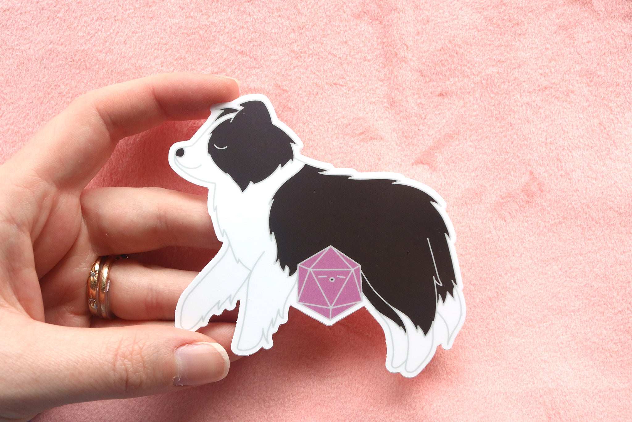 Hand holding a black and white border collie sticker. The sticker is of a sleepy puppy and mauve d20 dice.