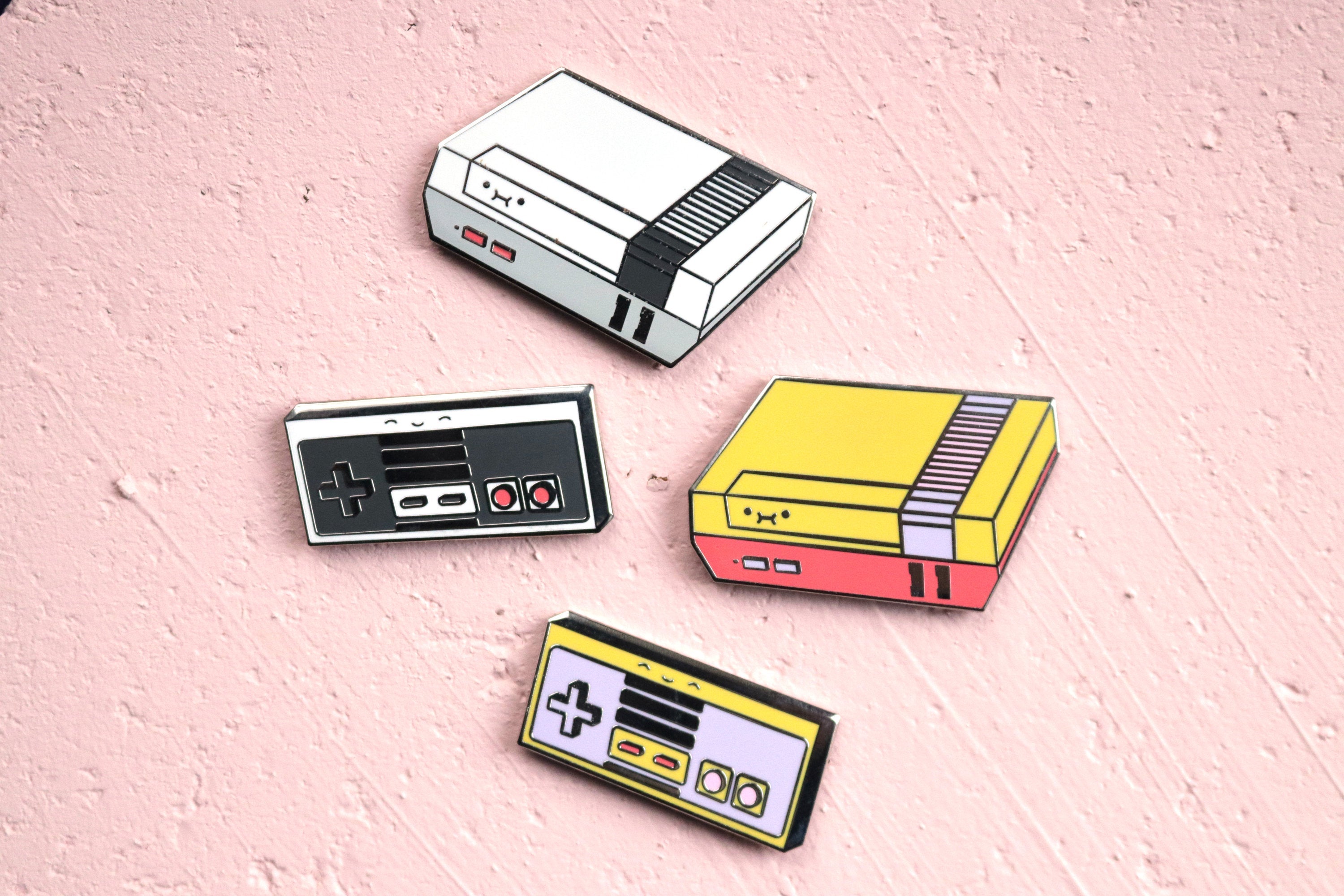 Gray NES Console and Controller with a Yellow NES Console and Controller on Pink Cork Board