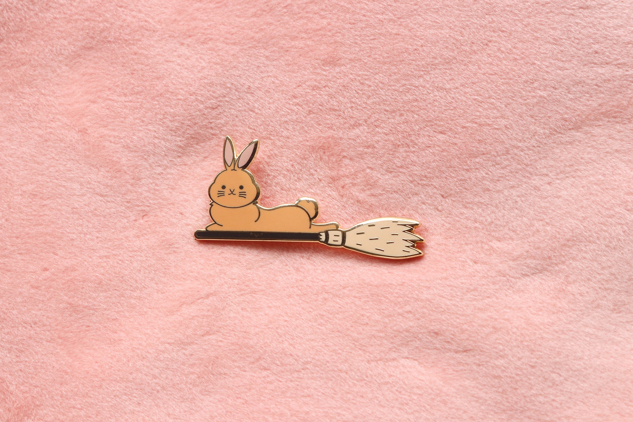 Enamel pin of an orange bunny riding a black broomstick.