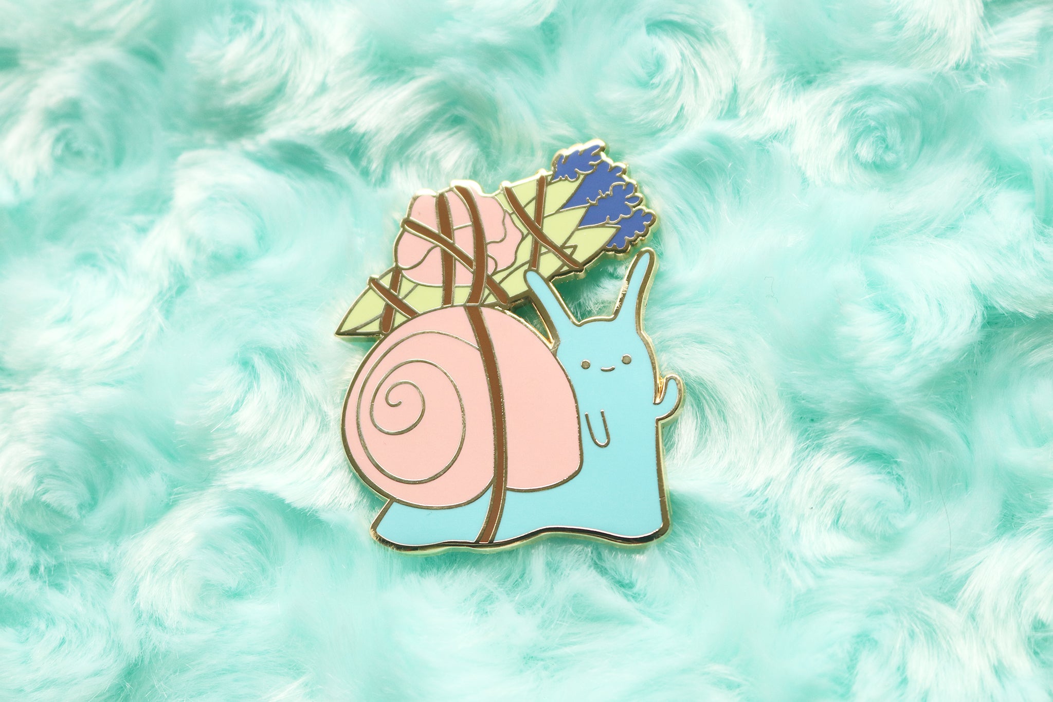 Enamel pin of a snail with a pink shell and blue body carrying a satchel of sage, rose petals, and lavender.