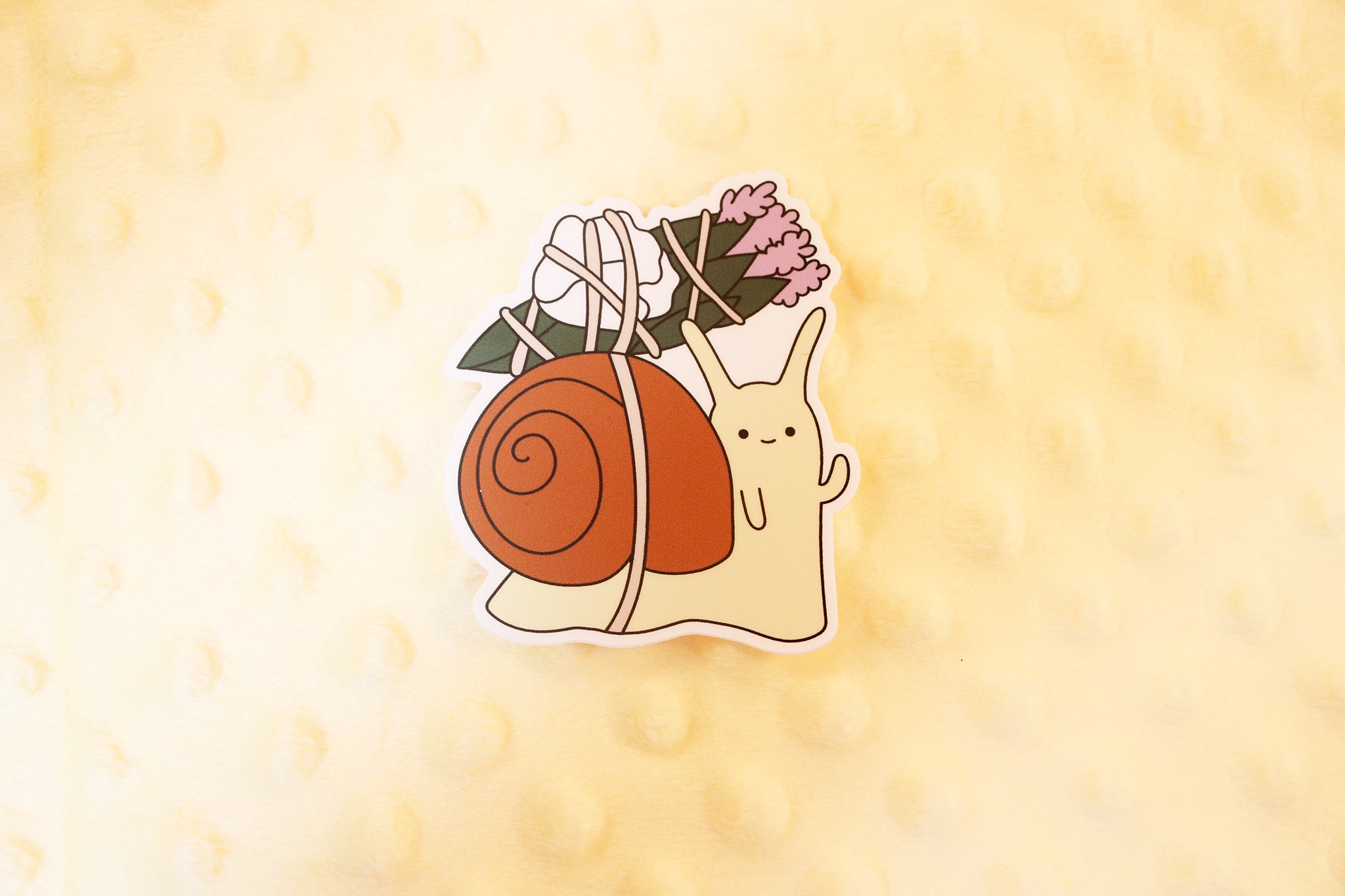 sticker of a snail with a brown shell and peige body carrying a satchel of sage, rose petals, and lavender.